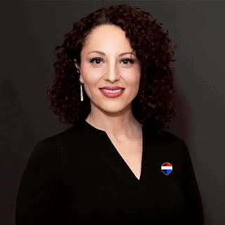 Professional headshot of a woman with curly dark red hair, wearing a black blazer and heart-shaped pin, smiling against a gray background