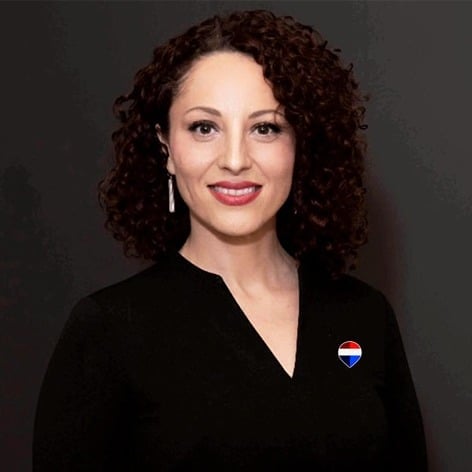 Professional headshot of a woman with curly dark red hair, wearing a black blazer and heart-shaped pin, smiling against a gray background